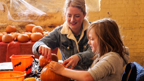 A parent and child carve pumpkins on a bench covered in pumpkins and orange buckets